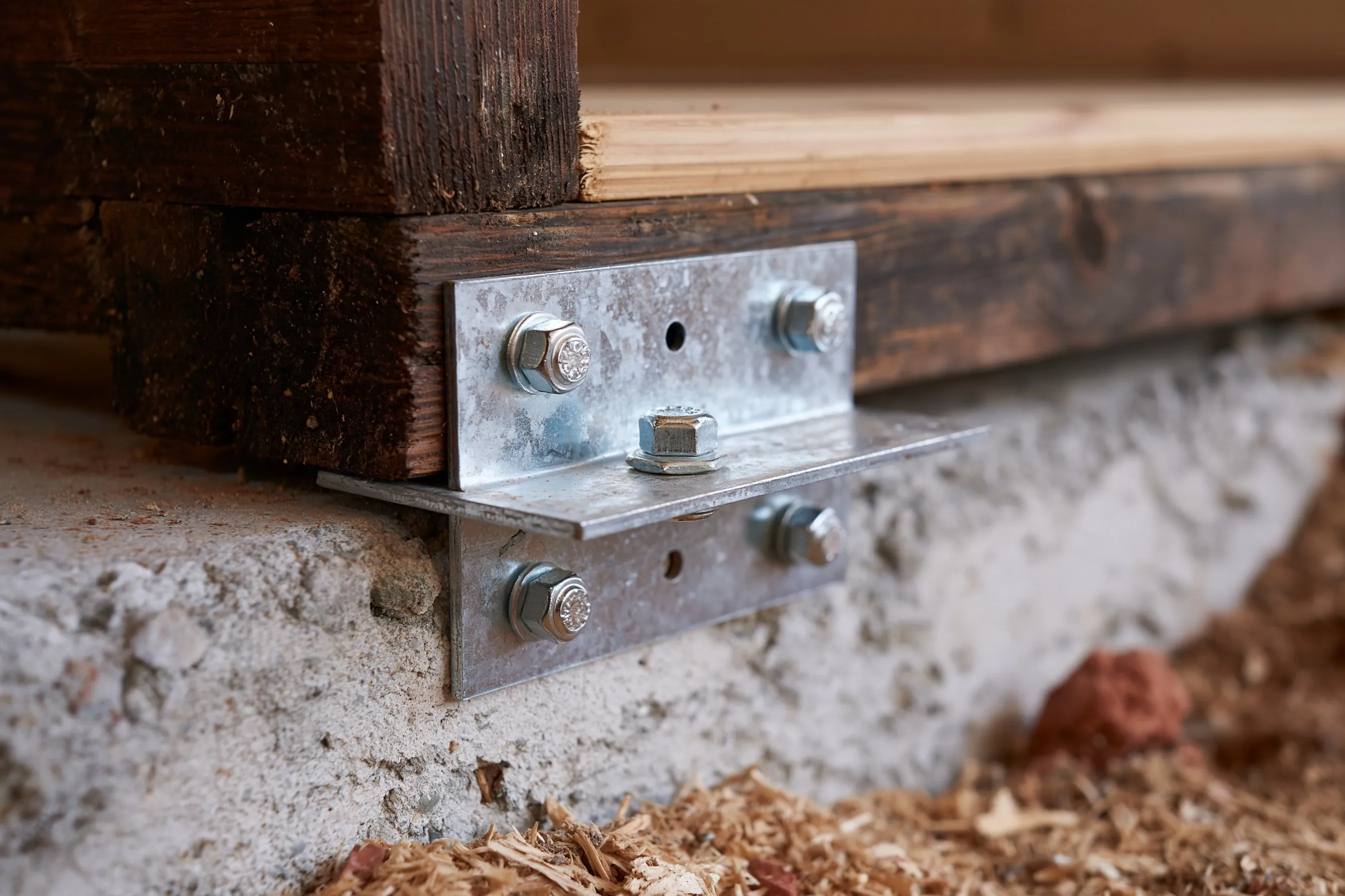 Galvanized steel connector plate fastening a floor joist and sill area to a concrete stem wall, with new bolts against older dark framing